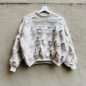 Könnte beinhalten: Ein cremefarbener Strickpullover mit Zopfmuster hängt an einem weißen Kleiderbügel vor einer beigefarbenen Wand.