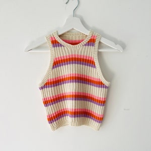 Puede incluir: Un top de tirantes blanco con rayas naranjas, rosas y moradas. El top es sin mangas y tiene un escote redondo.