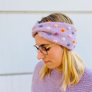 Op de afbeelding: Een lichtpaarse gebreide hoofdband met kleurrijke pompons. De hoofdband is gemaakt van een zacht, getextureerd garen en heeft een gedraaid ontwerp. De pompons zijn in tinten oranje, roze, blauw en paars. De hoofdband is ontworpen om de oren warm te houden.
