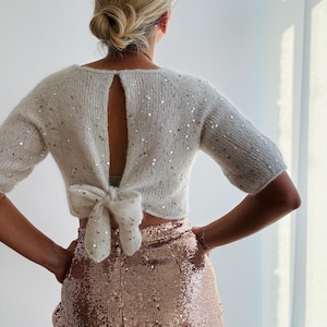 Può includere: Un top in maglia color crema a maniche corte con paillettes argentate, un'apertura verticale sulla schiena e un fiocco in vita, abbinato a una gonna a vita alta con paillettes color oro rosa che arriva sotto i fianchi.