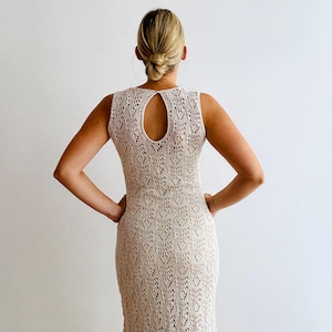 Peut inclure: Robe sans manches en crochet beige avec un dos en trou de serrure. La robe a un corsage ajusté et une jupe évasée. Le motif en crochet est complexe et détaillé, créant un aspect texturé. La robe est montrée de dos.