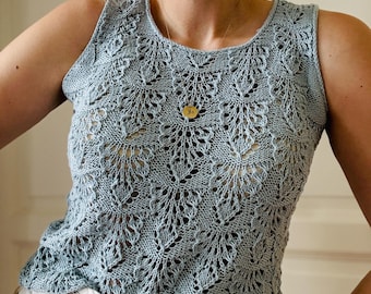 May include: A light blue, sleeveless knit top with a delicate, openwork floral pattern. The top has a scoop neckline and is paired with beige pants. A gold pendant hangs from a thin chain.