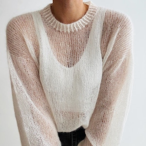 Puede incluir: Un suéter transparente blanco roto con mangas largas y cuello de canalé. El suéter está hecho de una tela delicada de punto abierto, que revela la prenda de abajo. El estilo es ligero y aireado, adecuado para superponer, con un ajuste relajado.