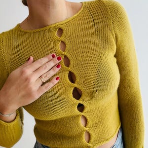 May include: Mustard yellow knit sweater with long sleeves and a round neckline. The sweater features a series of decorative, open oval cutouts down the front. The model is wearing blue jeans. The sweater is made of a soft, comfortable material.