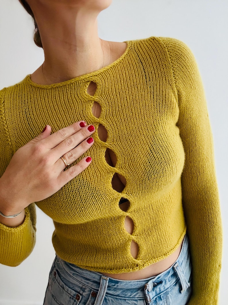 May include: Mustard yellow knit sweater with long sleeves and a round neckline. The sweater features a series of decorative, open oval cutouts down the front. The model is wearing blue jeans. The sweater is made of a soft, comfortable material.