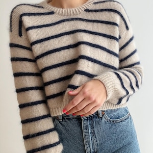 Puede incluir: Un suéter beige de cuello redondo con rayas horizontales azul marino. El suéter tiene mangas largas y se combina con unos vaqueros azules. El suéter está hecho de un material suave y texturizado.
