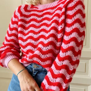 Peut inclure: Un pull rayé rose et rouge avec un motif ondulé. Le pull a un col rond et des manches longues.