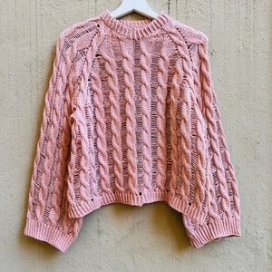 Könnte beinhalten: Ein rosa Strickpullover mit Zopfmuster, der an einem weißen Kleiderbügel an einer strukturierten Wand hängt.