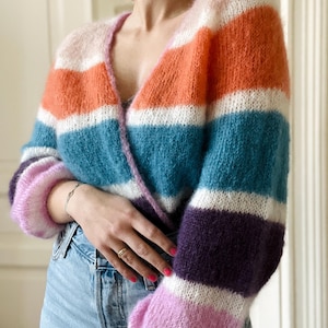 Op de afbeelding: Een veelkleurig gestreept vest in roze, oranje, blauw, wit en paars. De trui met lange mouwen heeft een V-hals en een zacht, getextureerd uiterlijk. Het vest wordt gecombineerd met een lichtblauwe spijkerbroek.