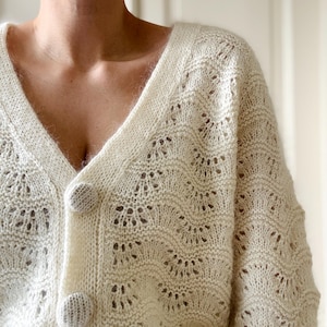 May include: A white crocheted cardigan with a V-neck and button closure. The cardigan has a delicate openwork pattern.