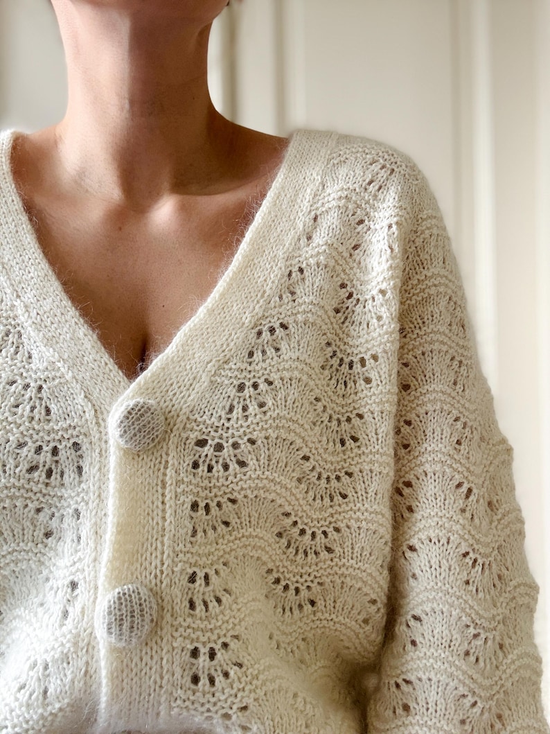 May include: A white crocheted cardigan with a V-neck and button closure. The cardigan has a delicate openwork pattern.