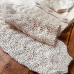 May include: A white knitted cardigan with a delicate openwork pattern. The cardigan is folded in half, showing the intricate details of the knit.