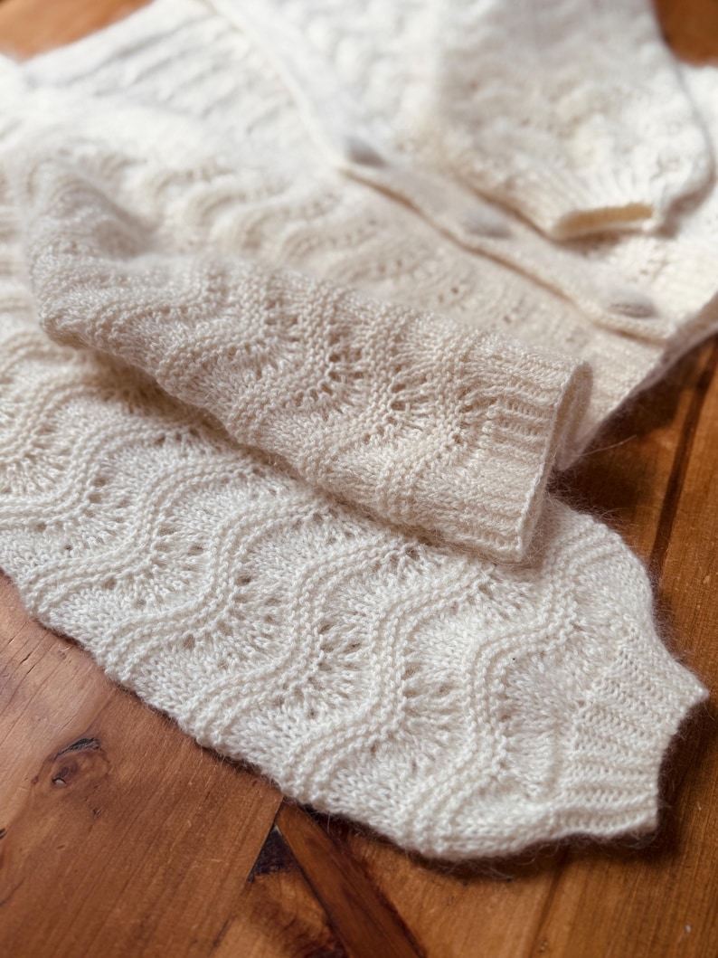 May include: A white knitted cardigan with a delicate openwork pattern. The cardigan is folded in half, showing the intricate details of the knit.