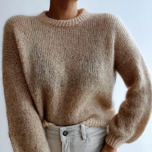 Könnte beinhalten: Ein beigefarbener Strickpullover mit Rundhalsausschnitt und langen Ärmeln. Der Pullover hat ein leicht schimmerndes Aussehen und wird mit hellen Hosen kombiniert. Der Gesamtstil ist lässig und bequem.