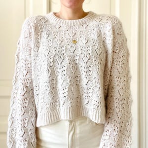 May include: A white crocheted sweater with a delicate openwork pattern. The sweater has a cropped length and long sleeves.