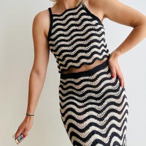 Könnte beinhalten: Zweiteiliges Häkelset in Schwarz und Creme. Das Oberteil ist ein Neckholder-Modell mit gewelltem Saum, der Rock ist ein eng anliegendes Midikleid. Das Wellenmuster verleiht dem Outfit eine einzigartige Textur. Das Set ist aus leichtem Material gefertigt.
