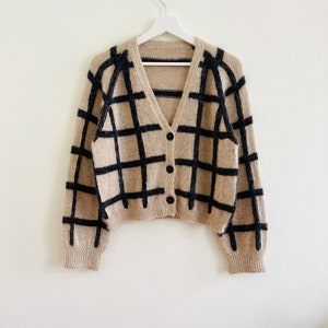 Könnte beinhalten: Beige Cardigan mit schwarzem Gittermuster, Knopfverschluss und gerippten Bündchen. Der Pullover hängt an einem weißen Kleiderbügel vor weißem Hintergrund. Der Cardigan ist im V-Ausschnitt-Stil.