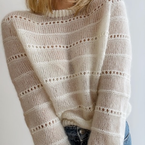 Könnte beinhalten: Ein cremefarbener, offen gestrickter Pullover mit horizontalen Streifendetails. Der Pullover hat einen Rundhalsausschnitt und lange Ärmel. Das Strickmuster enthält kleine, gleichmäßig verteilte Löcher, die eine zarte Textur erzeugen. Das Kleidungsstück ist leicht und wirkt leicht durchsichtig.