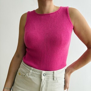 May include: A vibrant pink tank top with a ribbed knit texture. The top has a scoop neckline and is sleeveless. The model is wearing light-colored jeans. The top is a solid color and appears to be made of a soft material.