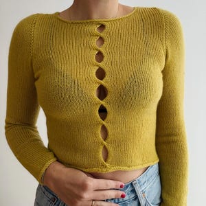 May include: Mustard yellow long-sleeved knit top with a scoop neckline and a series of oval-shaped openings down the center. The top is cropped and has ribbed detailing at the cuffs and hem.