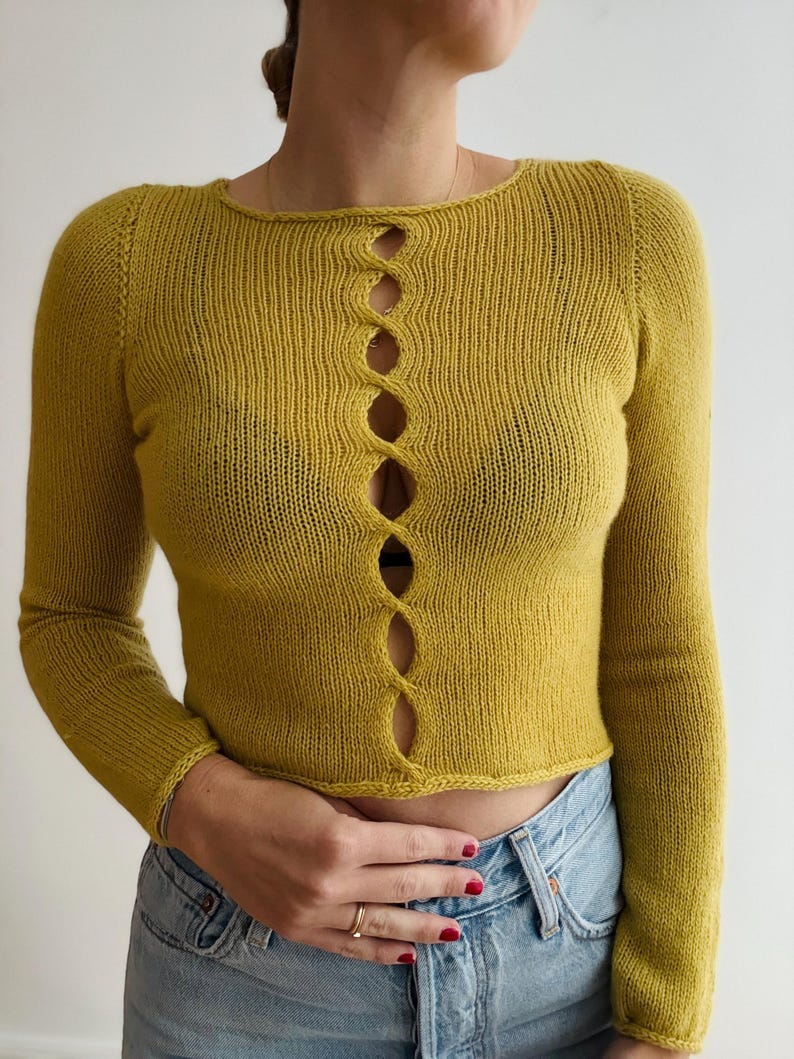 May include: Mustard yellow long-sleeved knit top with a scoop neckline and a series of oval-shaped openings down the center. The top is cropped and has ribbed detailing at the cuffs and hem.