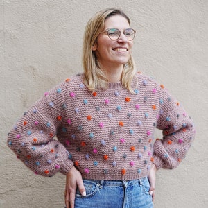 Könnte beinhalten: Eine Frau trägt einen rosa Pullover mit bunten Punkten und blaue Jeans. Sie lächelt und schaut nach rechts.