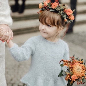 Puede incluir: Una niña pequeña con un suéter de punto azul claro con un dobladillo con volantes y una falda floral. Lleva un ramo de flores naranjas y amarillas y una corona de flores con flores naranjas y rojas.