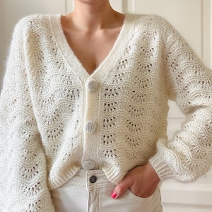 Könnte beinhalten: Ein weißer Häkelcardigan mit V-Ausschnitt und Knopfleiste. Der Cardigan hat ein zartes Lochmuster und eine leicht verkürzte Länge.