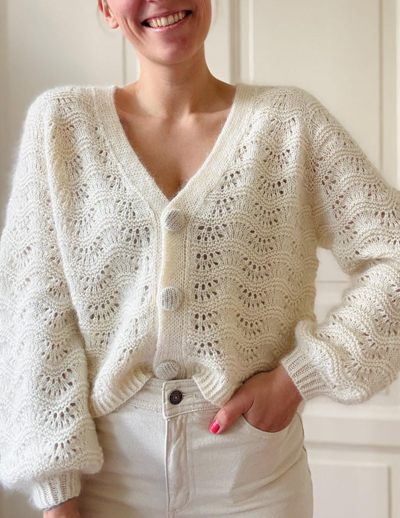 May include: A white crocheted cardigan with a V-neck and button closure. The cardigan has a delicate openwork pattern and a slightly cropped length.