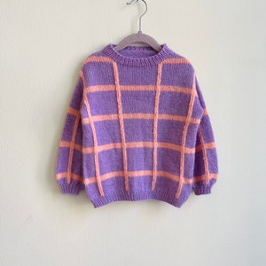 Può includere: Un maglione di maglia a quadri viola e arancio appeso a un appendiabiti di legno su uno sfondo bianco.