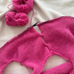 May include: A work-in-progress knitting project featuring vibrant pink yarn and knitting needles. Two balls of yarn sit beside the partially completed knitted fabric. The fabric has two openings, suggesting a garment is being made.