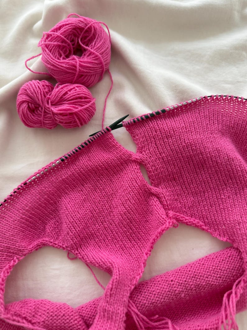 May include: A work-in-progress knitting project featuring vibrant pink yarn and knitting needles. Two balls of yarn sit beside the partially completed knitted fabric. The fabric has two openings, suggesting a garment is being made.