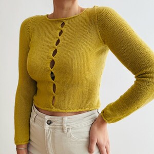 May include: Mustard yellow long-sleeved cardigan with a round neckline and a series of oval-shaped openings down the front. The cardigan is made of a knitted material and is paired with light-colored pants.
