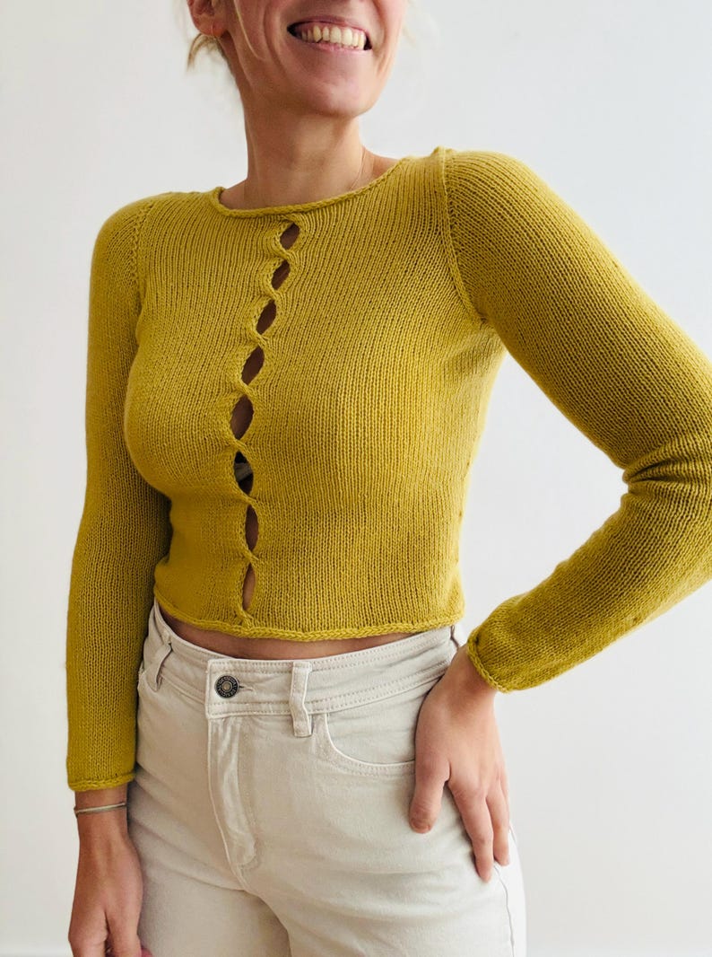 May include: Mustard yellow long-sleeved cardigan with a round neckline and a series of oval-shaped openings down the front. The cardigan is made of a knitted material and is paired with light-colored pants.
