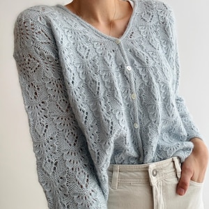 Op de afbeelding: Een lichtblauwe, opengebreide cardigan met een V-hals en knoopsluiting. De cardigan heeft een delicaat bloemmotief en wordt gecombineerd met een lichtgekleurde broek. De mouwen zijn lang en de algehele stijl is zacht en vrouwelijk.