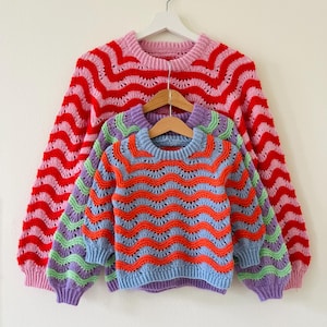 Könnte beinhalten: Drei gestreifte Pullover hängen an einem weißen Kleiderbügel. Der oberste Pullover ist rosa und rot, der mittlere Pullover ist grün und blau und der unterste Pullover ist lila und orange.