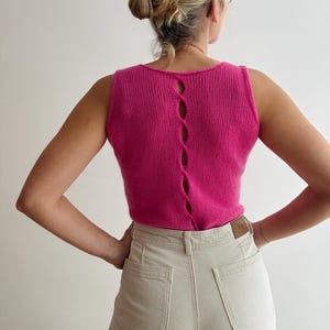 May include: A sleeveless, hot pink knit top with a scoop neck and a vertical, open back design. The back features four oval-shaped openings. The top is paired with light beige jeans.