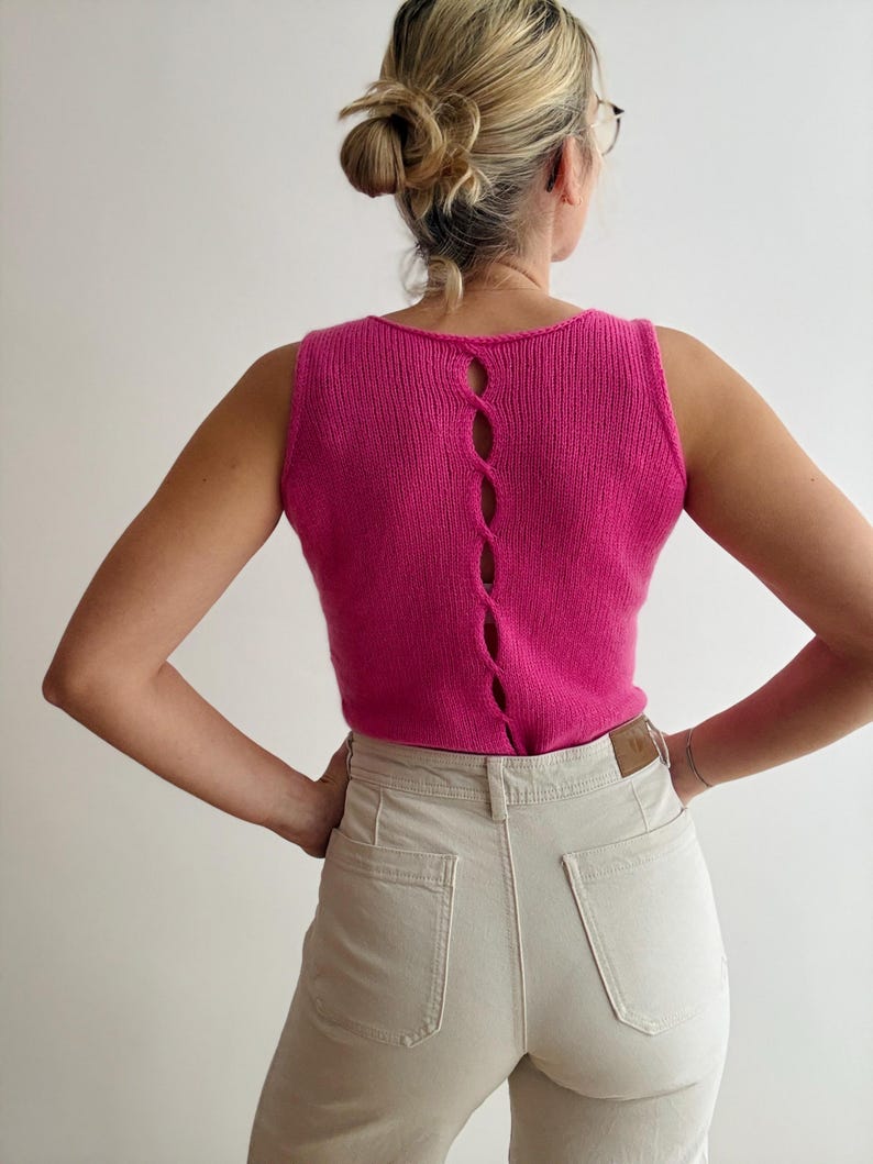 May include: A sleeveless, hot pink knit top with a scoop neck and a vertical, open back design. The back features four oval-shaped openings. The top is paired with light beige jeans.