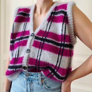 May include: A sleeveless knitted waistcoat with a plaid pattern in shades of pink, grey, and black. The waistcoat has a V-neck and button closure.