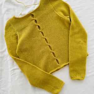 May include: Mustard yellow knitted cardigan with long sleeves and a unique button-hole design down the front. The sweater is displayed on a gold hanger against a white background. The cardigan is a soft, textured knit.