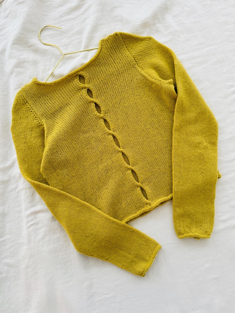 May include: Mustard yellow knitted cardigan with long sleeves and a unique button-hole design down the front. The sweater is displayed on a gold hanger against a white background. The cardigan is a soft, textured knit.