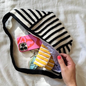 Könnte beinhalten: Eine gehäkelte Tasche mit schwarzen und weißen Streifen und einem Reißverschluss. Die Tasche ist offen und enthält eine rosa Handyhülle, eine gelbe und weiße gestreifte Tasche und einen Schlüsselanhänger.