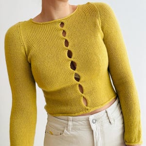 May include: Mustard yellow knit cardigan with long sleeves and a round neckline. The cardigan features a series of vertical openings down the front. The model is wearing light-colored jeans. The cardigan is a stylish addition to any wardrobe.
