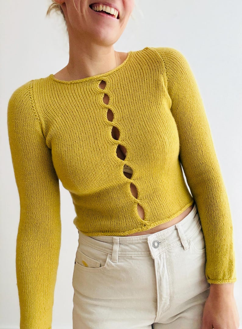 May include: Mustard yellow knit cardigan with long sleeves and a round neckline. The cardigan features a series of vertical openings down the front. The model is wearing light-colored jeans. The cardigan is a stylish addition to any wardrobe.