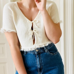 Könnte beinhalten: Cremefarbenes, kurzärmeliges Strickoberteil mit V-Ausschnitt und zarten Schleifen. Das Oberteil hat einen gerüschten Saum und Ärmelränder. Kombiniert mit einer hoch taillierten Jeans aus blauem Denim.
