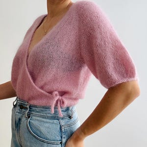Könnte beinhalten: Ein hellrosa Wickelpullover mit kurzen Ärmeln und einem Bindeband. Der Pullover besteht aus einem weichen, strukturierten Material und wird mit hellblauen Jeans kombiniert.