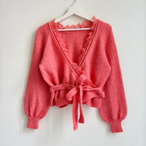 以下が含まれることがあります： レースのトリムネックラインとタイベルトが付いたコーラルピンクのニットカーディガン。カーディガンは長袖で、ラップスタイルです。