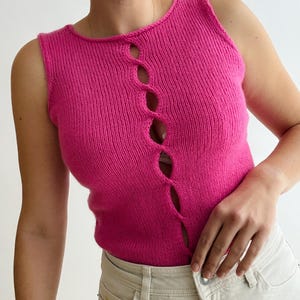 May include: A sleeveless, hot pink knit top with a unique cutout design down the center. The top is made of a soft, textured material and has a scoop neckline. The model is wearing the top with light-colored jeans.