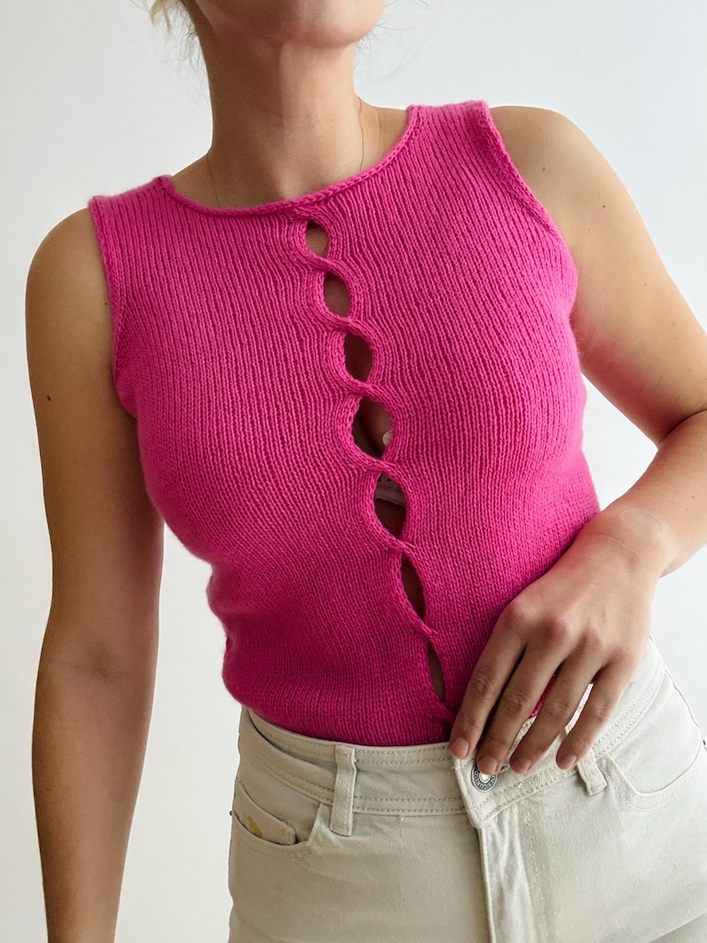 May include: A sleeveless, hot pink knit top with a unique cutout design down the center. The top is made of a soft, textured material and has a scoop neckline. The model is wearing the top with light-colored jeans.