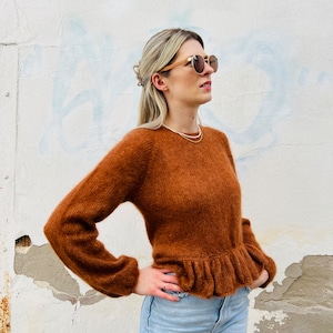 May include: A brown fuzzy sweater with a ruffled hem. The sweater has long sleeves and a round neckline.